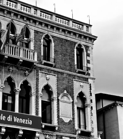 Czarno-biała fotografia. Budynek w Wenecji. Na balustradzie zawieszony baner z napisem La Biennale di Venezia.