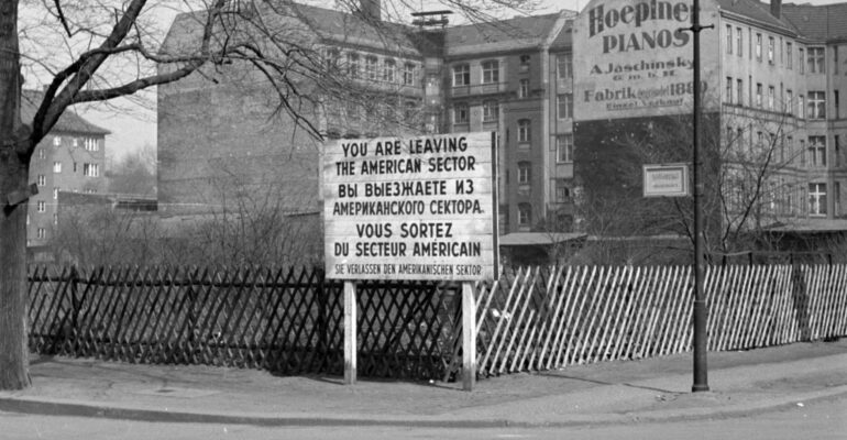 Czarno-biała fotografia. Fragment Berlina Zachodniego. Na ulicy stoi tablica informująca w trzech językach (angielskim, niemieckim i rosyjskim) o opuszczeniu sektora amerykańskiego.