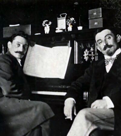 Czarno-biała fotografia. Dwóch elegancko ubranych mężczyzn siedzi przy fortepianie. Spoglądają w stronę fotografa.