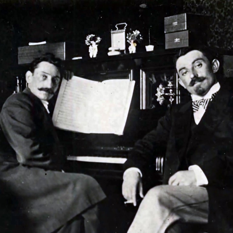 Czarno-biała fotografia. Dwóch elegancko ubranych mężczyzn siedzi przy fortepianie. Spoglądają w stronę fotografa.