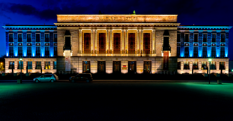Pałac Kultury Zagłębia. Noc. Fasada budynku oświetlona jest kolorowymi lampami. Przed budynkiem zaparkowane dwa samochody.