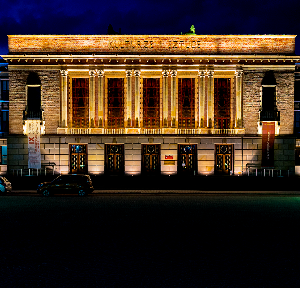 Pałac Kultury Zagłębia. Noc. Fasada budynku oświetlona jest kolorowymi lampami. Przed budynkiem zaparkowane dwa samochody.