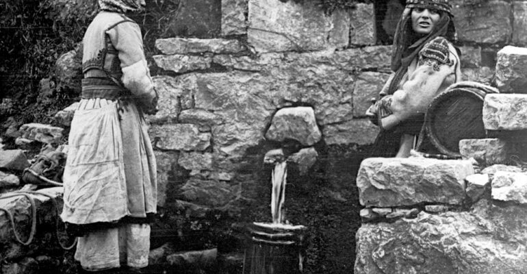 Obrazek czarnobiały. Dwie kobiety siedzą przy kamiennej ścianie. Na ziemi stoi wiadro, do którego wpada woda.