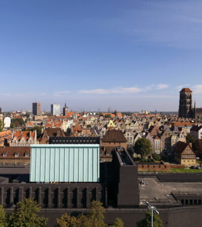 Panorama historycznego centrum Gdańska oglądana z góry w słoneczny dzień. Na pierwszym planie widoczny jest nowoczesny budynek Gdańskiego Teatru Szekspirowskiego o ciemnej bryle i jasnej, geometrycznej części centralnej. W tle rozciąga się gęsta zabudowa Starego i Głównego Miasta z charakterystycznymi ceglastymi dachami oraz licznymi zabytkowymi kamienicami. Po prawej stronie dominuje monumentalna sylwetka Bazyliki Mariackiej. Nad miastem rozciąga się bezchmurne, błękitne niebo.