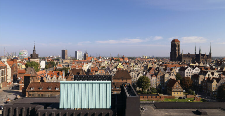 Panorama historycznego centrum Gdańska oglądana z góry w słoneczny dzień. Na pierwszym planie widoczny jest nowoczesny budynek Gdańskiego Teatru Szekspirowskiego o ciemnej bryle i jasnej, geometrycznej części centralnej. W tle rozciąga się gęsta zabudowa Starego i Głównego Miasta z charakterystycznymi ceglastymi dachami oraz licznymi zabytkowymi kamienicami. Po prawej stronie dominuje monumentalna sylwetka Bazyliki Mariackiej. Nad miastem rozciąga się bezchmurne, błękitne niebo.