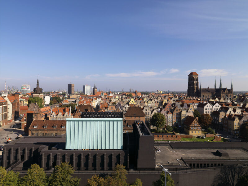 Panorama historycznego centrum Gdańska oglądana z góry w słoneczny dzień. Na pierwszym planie widoczny jest nowoczesny budynek Gdańskiego Teatru Szekspirowskiego o ciemnej bryle i jasnej, geometrycznej części centralnej. W tle rozciąga się gęsta zabudowa Starego i Głównego Miasta z charakterystycznymi ceglastymi dachami oraz licznymi zabytkowymi kamienicami. Po prawej stronie dominuje monumentalna sylwetka Bazyliki Mariackiej. Nad miastem rozciąga się bezchmurne, błękitne niebo.