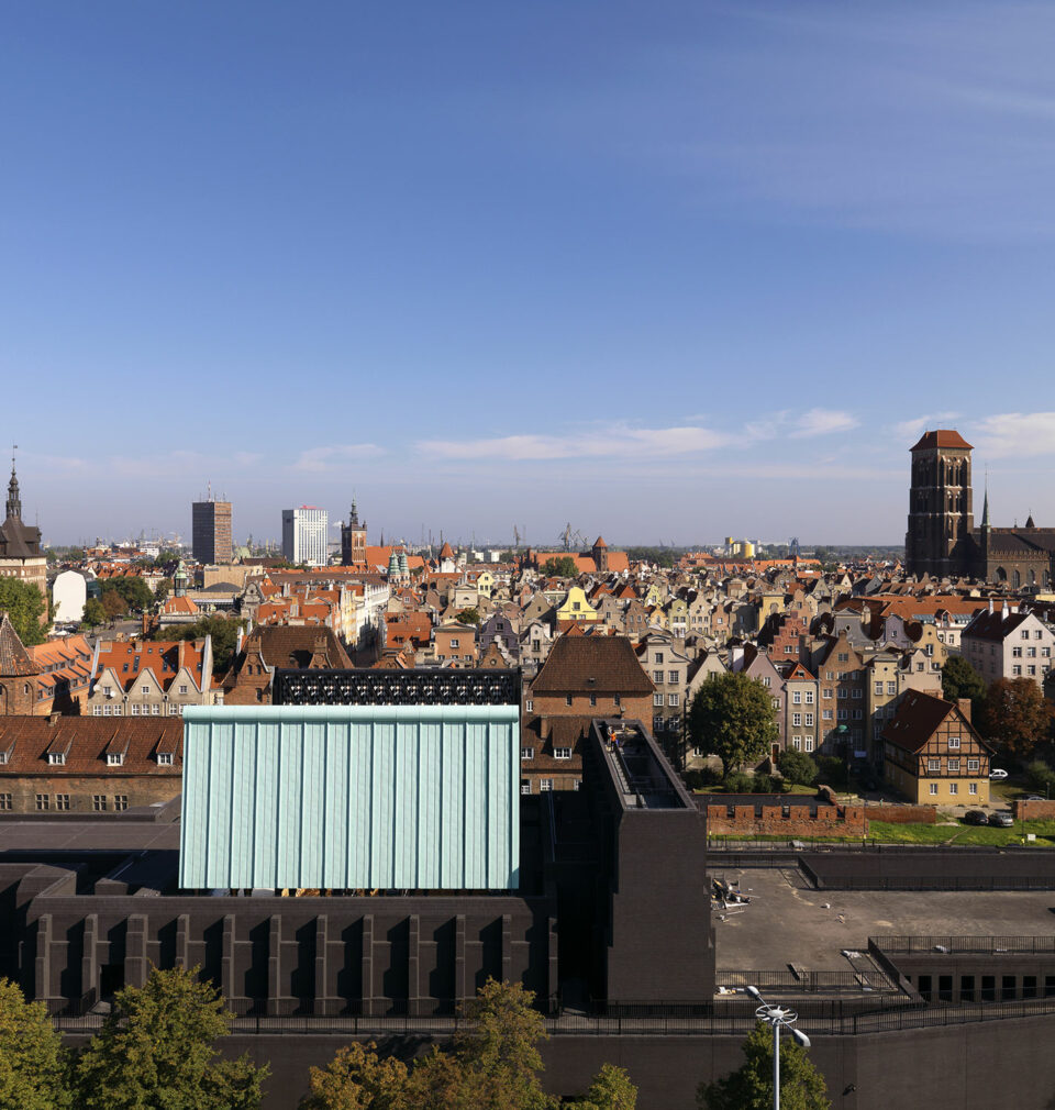 Panorama historycznego centrum Gdańska oglądana z góry w słoneczny dzień. Na pierwszym planie widoczny jest nowoczesny budynek Gdańskiego Teatru Szekspirowskiego o ciemnej bryle i jasnej, geometrycznej części centralnej. W tle rozciąga się gęsta zabudowa Starego i Głównego Miasta z charakterystycznymi ceglastymi dachami oraz licznymi zabytkowymi kamienicami. Po prawej stronie dominuje monumentalna sylwetka Bazyliki Mariackiej. Nad miastem rozciąga się bezchmurne, błękitne niebo.
